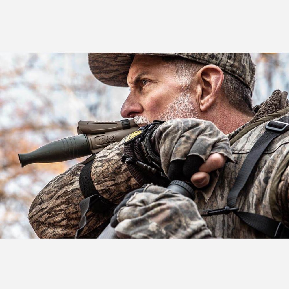 Man in camouflage holding a rifle with a scope, outdoors.