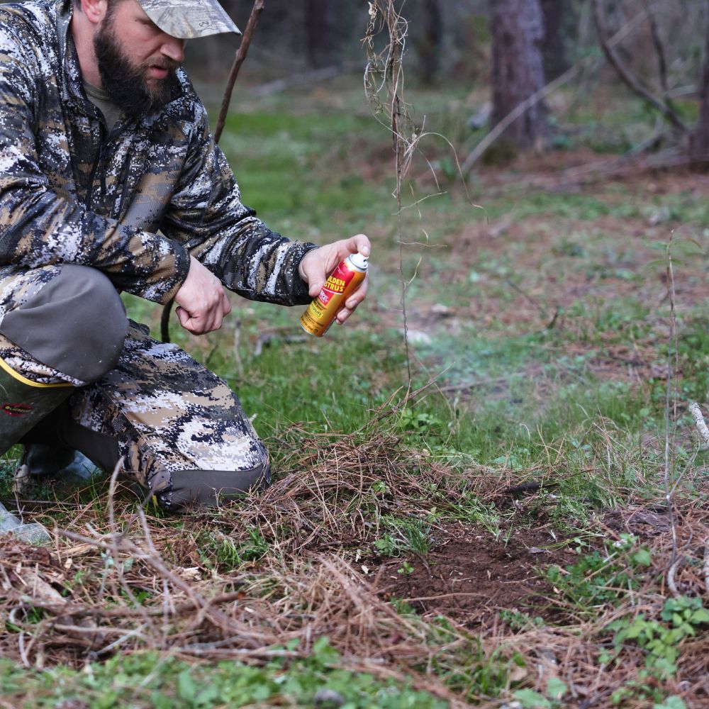 Man in camouflage clothing applying a spray can to the ground in a forest setting