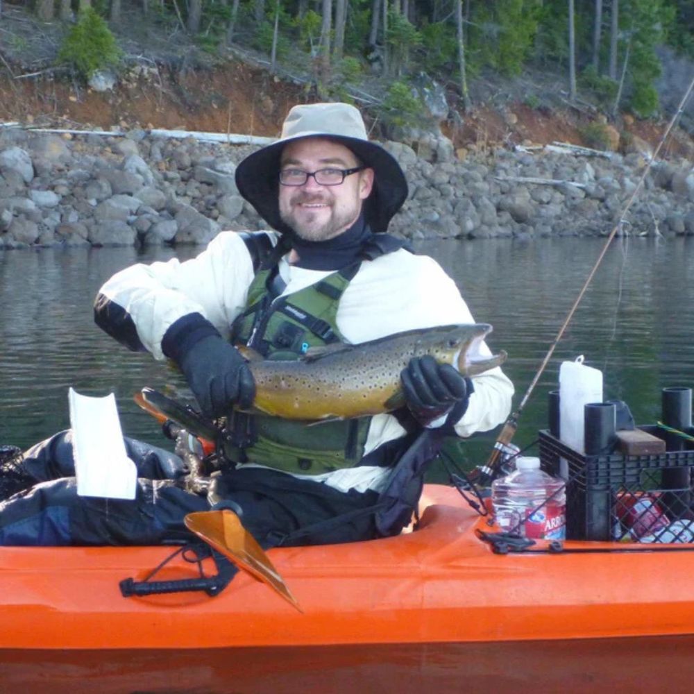 Man in kayak holding a fish on a lake with trees and rocks in the background