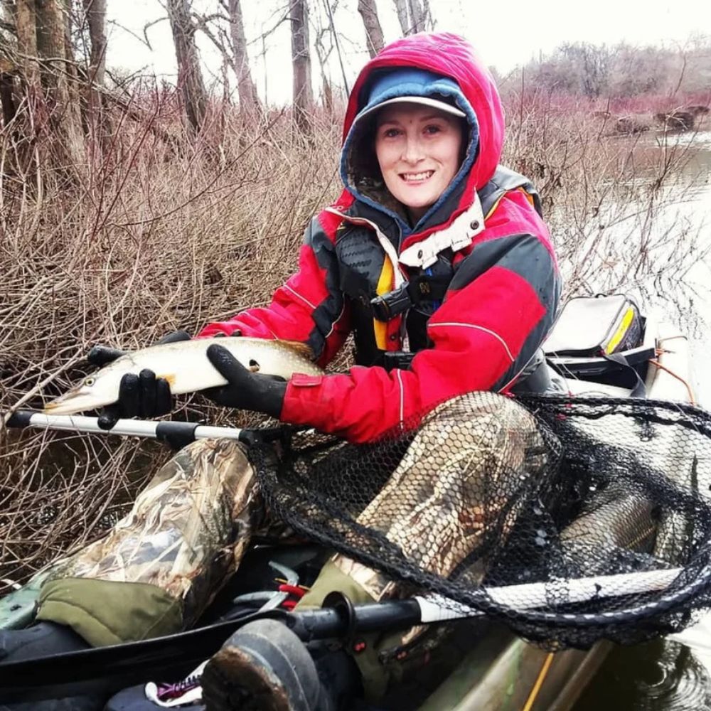 Person holding a fish in a kayak on a riverbank with trees and water in the background