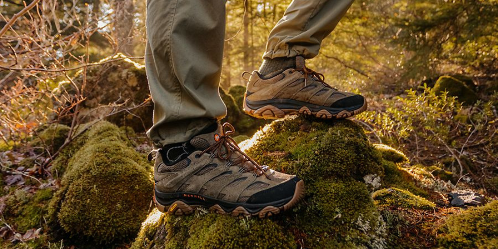 Hiker stepping across mossy rocks on a forest trail wearing rugged Merrell hiking shoes