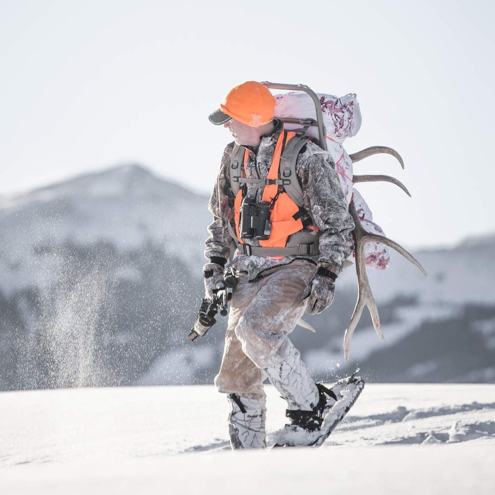 Hunter in winter gear with a backpack and skis, standing on snow with mountains in the background.