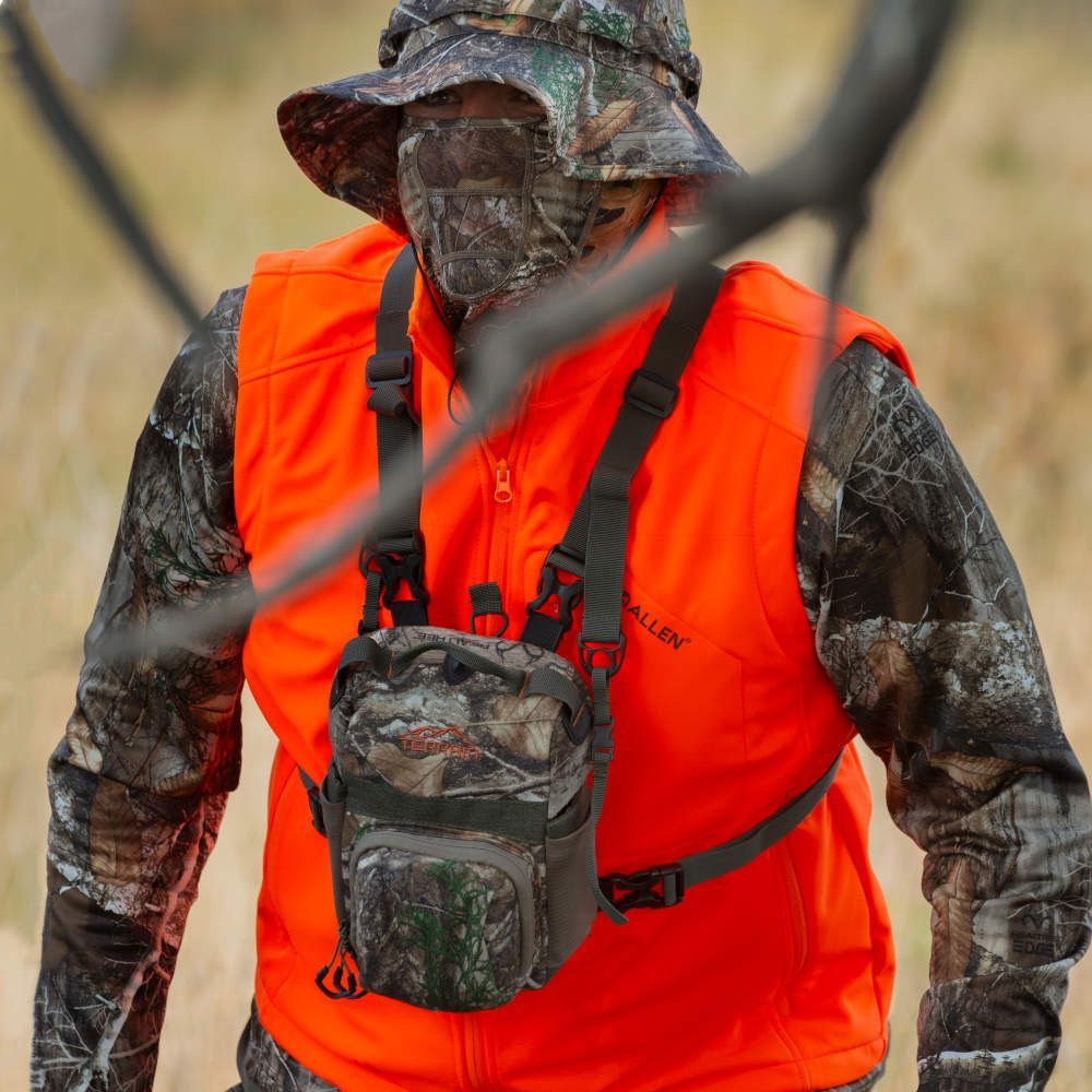 Person in camouflage clothing and an orange vest with a small bag, standing in a natural setting.