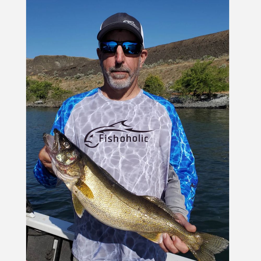 A man on a boat wearing a Fishoholic brand fishing shirt holds a large fish. The shirt features a white front with a blue water-texture pattern on the sleeves and sides, with a black Fishoholic logo printed on the chest.