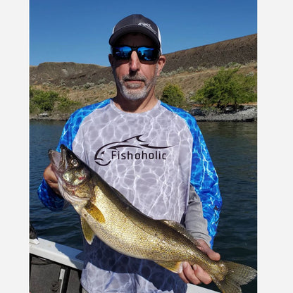 A man on a boat wearing a Fishoholic brand fishing shirt holds a large fish. The shirt features a white front with a blue water-texture pattern on the sleeves and sides, with a black Fishoholic logo printed on the chest.