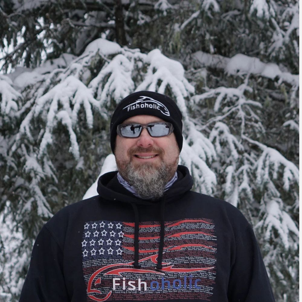 A man wearing a black beanie and a black hoodie with a red and white American flag graphic and 'Fishaholic' logo, standing in front of snow-covered pine trees.