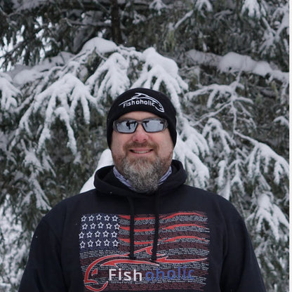 A man wearing a black beanie and a black hoodie with a red and white American flag graphic and 'Fishaholic' logo, standing in front of snow-covered pine trees.