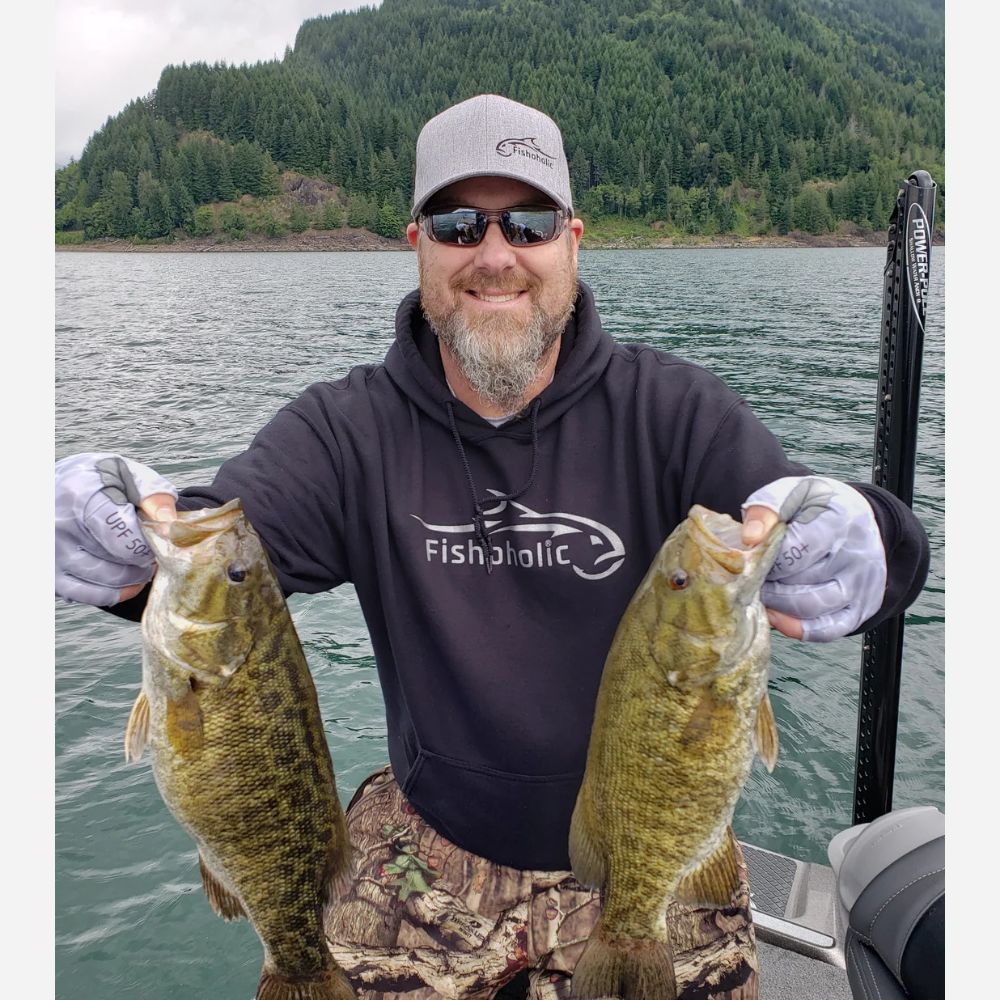 A man wearing a gray cap, sunglasses, and a black 'Fishalotic' hoodie holds two largemouth bass on a boat with a forested mountain in the background.