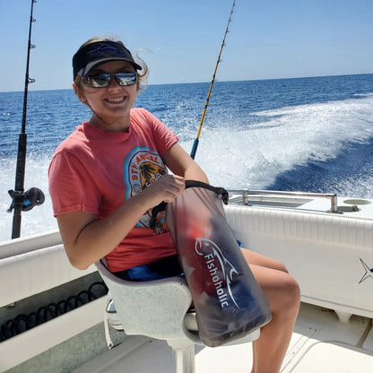 A woman sitting on a boat seat holding a translucent waterproof pouch branded 'Fisholitic' with blue and red items inside, with ocean water and fishing rods in the background.