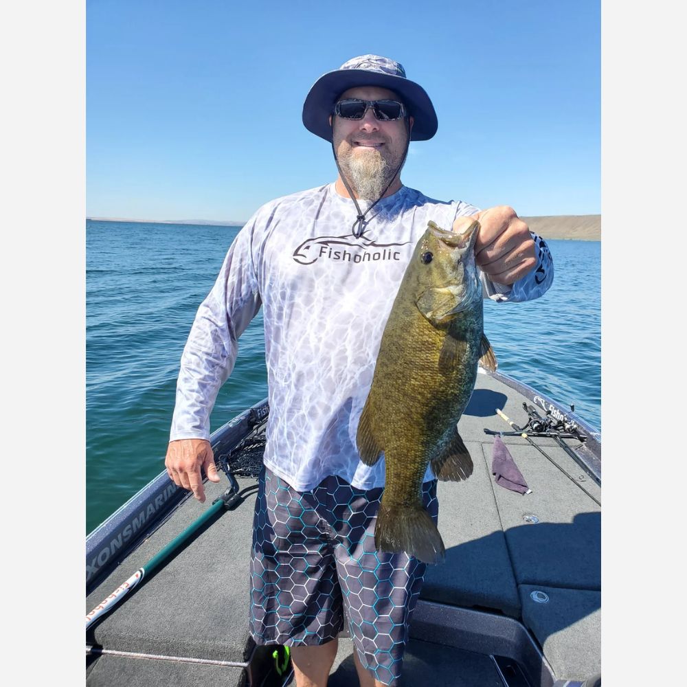 A man wearing a Fishingnole long-sleeve shirt and patterned shorts stands on a boat holding a largemouth bass under a clear blue sky.