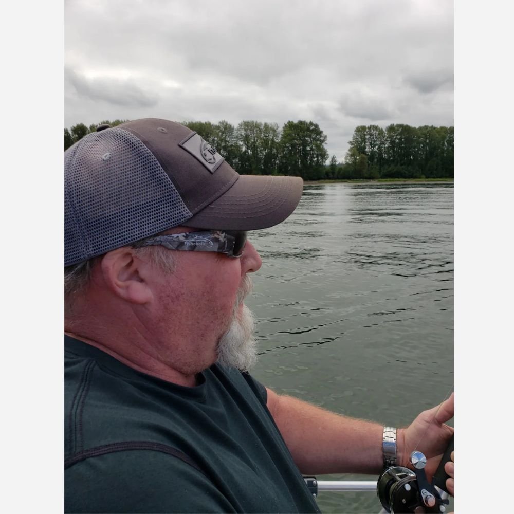 A man wearing a dark gray mesh-back baseball cap, sunglasses, and a dark green t-shirt sitting on a boat on the water, looking out towards a tree-lined shore under a cloudy sky.