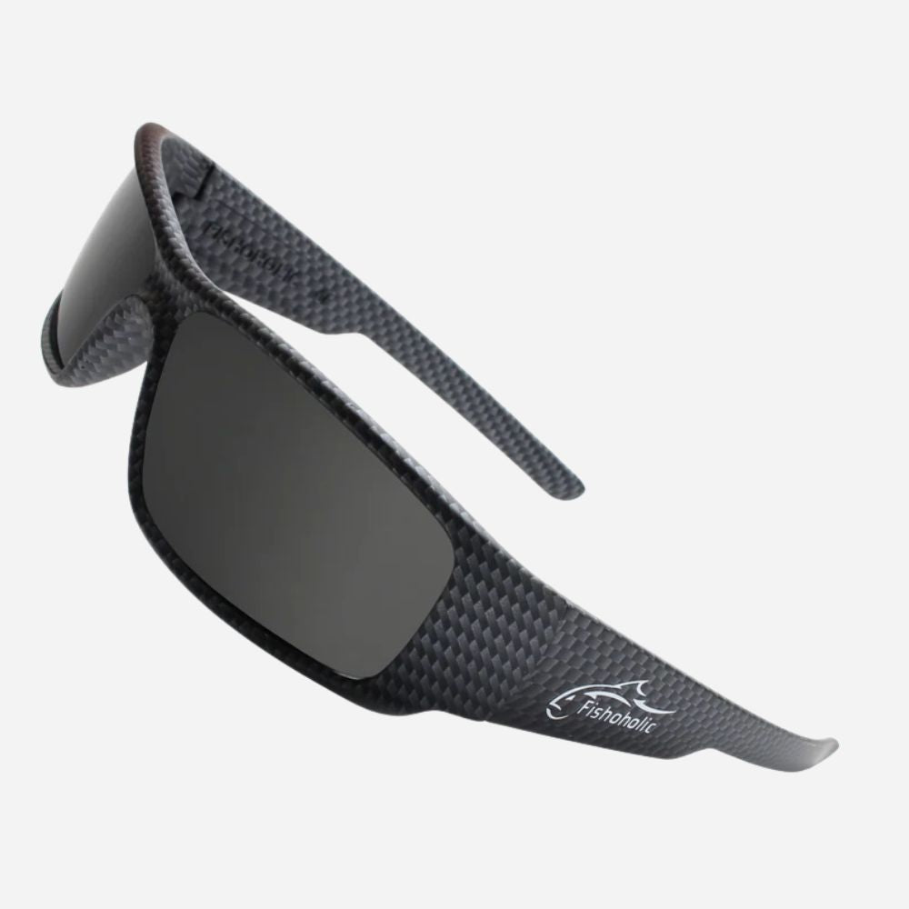 A pair of wrap-around sunglasses with a dark gray carbon fiber textured frame and dark lenses, featuring the Fairbairn logo on the temple, set against a light gray background.