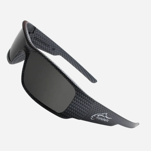 A pair of wrap-around sunglasses with a dark gray carbon fiber textured frame and dark lenses, featuring the Fairbairn logo on the temple, set against a light gray background.