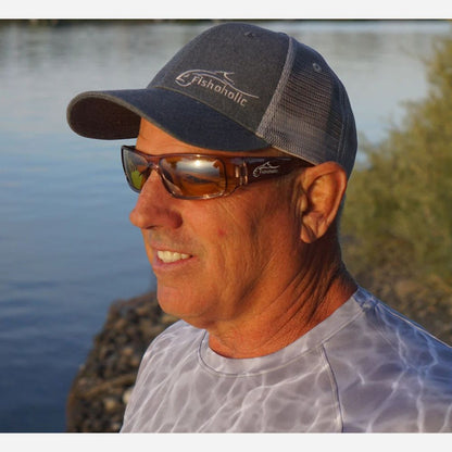 A man wearing a dark gray Fishholic trucker hat and brown-tinted sunglasses, photographed outdoors near water with a warm golden light.