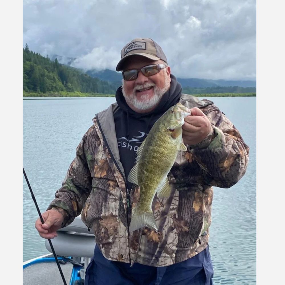 A man wearing a camouflage hunting jacket, black hoodie, and cap holds a freshly caught largemouth bass on a boat with a lake and forested mountains in the background.