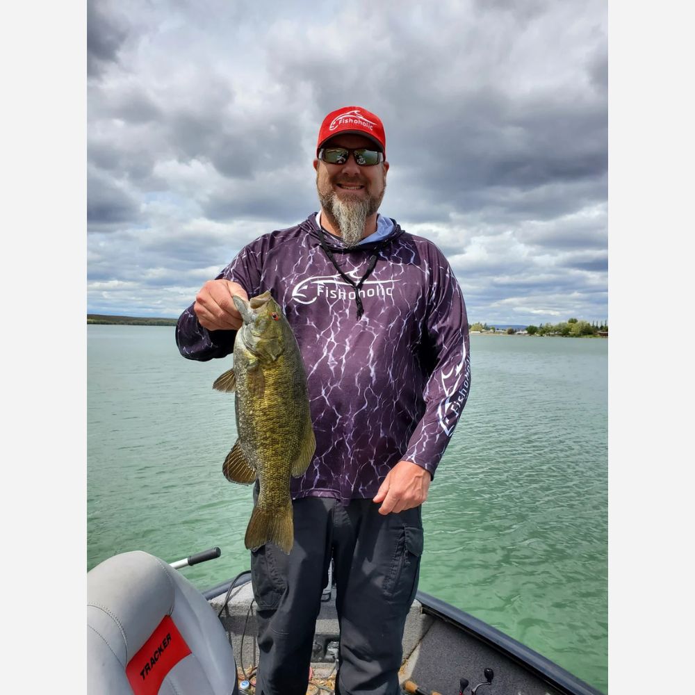 A bearded man wearing a red cap, sunglasses, and a purple patterned long-sleeve fishing shirt stands on a boat holding a largemouth bass, with water and a cloudy sky in the background.
