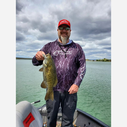 A bearded man wearing a red cap, sunglasses, and a purple patterned long-sleeve fishing shirt stands on a boat holding a largemouth bass, with water and a cloudy sky in the background.