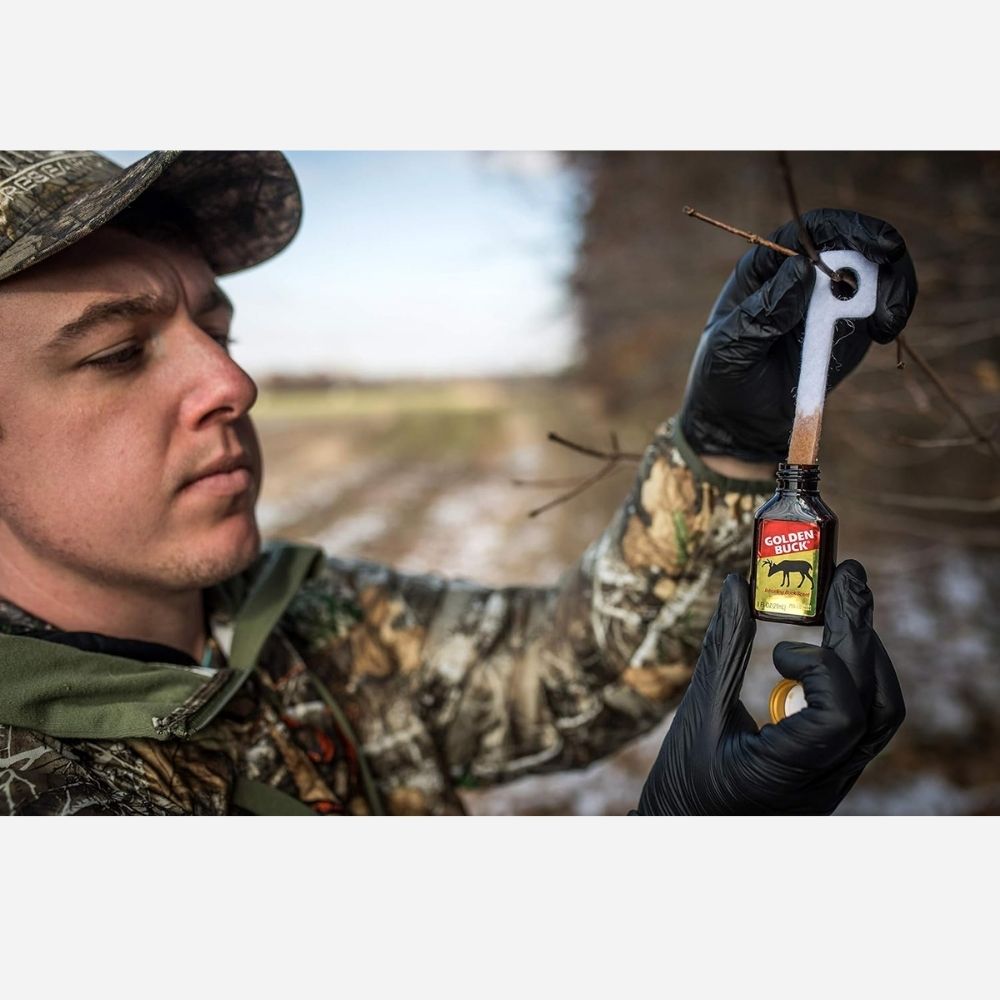 Person wearing camouflage holding Golden Buck scent attractant bottle and lifting a scent wick outdoors
