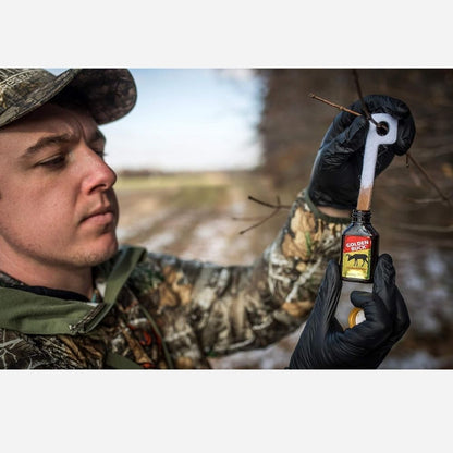 Person wearing camouflage holding Golden Buck scent attractant bottle and lifting a scent wick outdoors