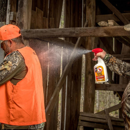 Person in orange vest and cap using a spray bottle on a horse in a wooden stable.