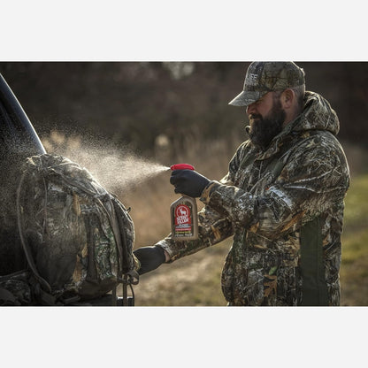 Man in camouflage gear applying a product from a bottle to a backpack outdoors.