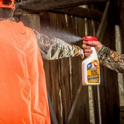 Person spraying scent control spray onto camouflage clothing with orange safety vest outdoors