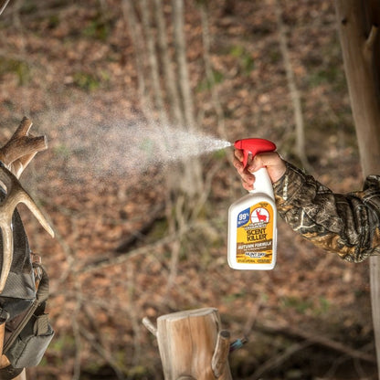 Person spraying scent control spray from a bottle outdoors near hunting gear