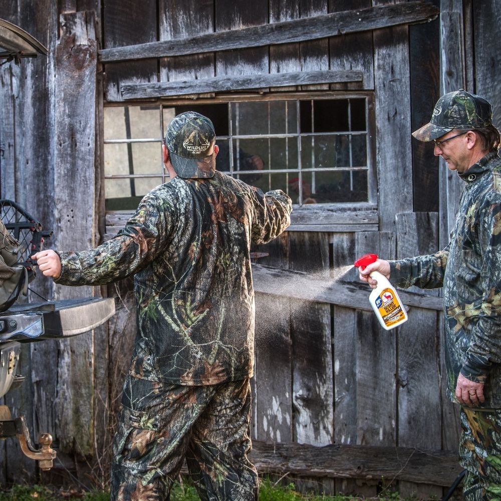 Person spraying scent control spray onto camouflage clothing worn by another person outdoors near a wooden building