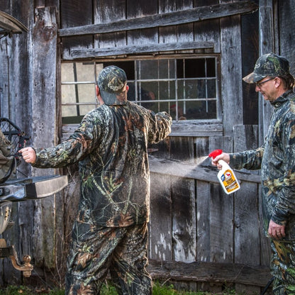 Person spraying scent control spray onto camouflage clothing worn by another person outdoors near a wooden building