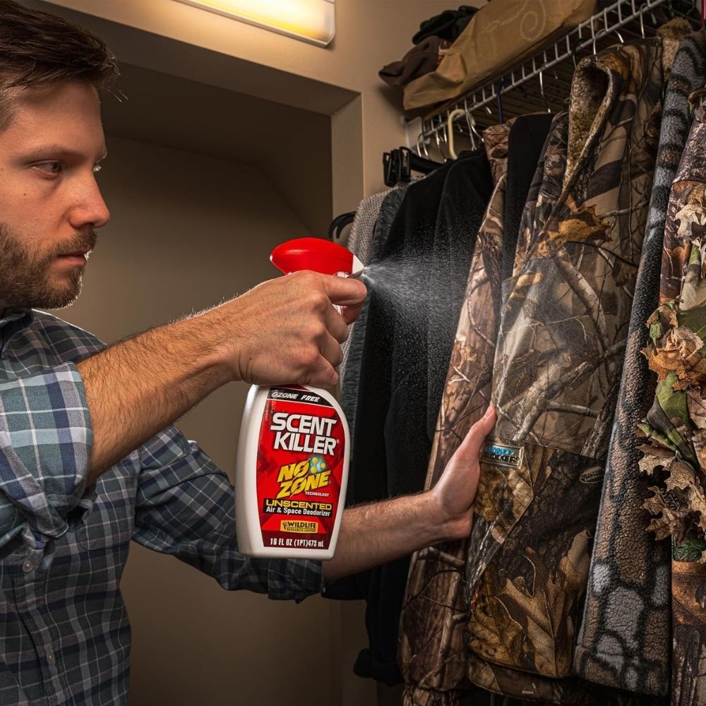 Person spraying Scent Killer No Zone deodorizer onto camouflage clothing hanging in a closet