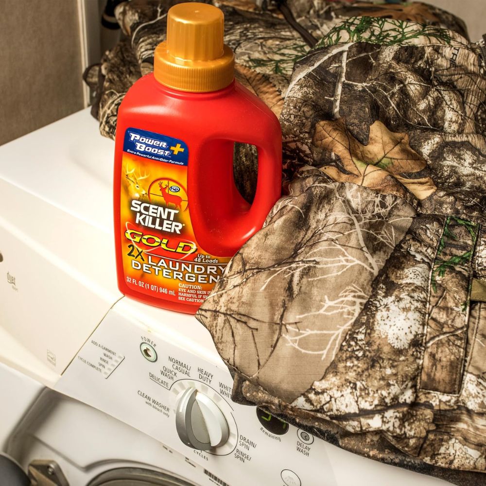 Red bottle of Power Bright Scent Killer Gold laundry detergent on a camouflage-patterned fabric with a washing machine in the background.
