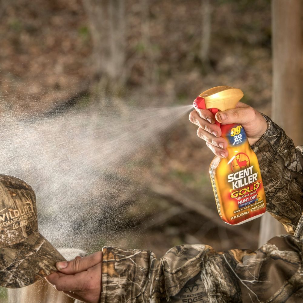 Person in camouflage clothing using a Scent Killer Gold spray bottle outdoors.