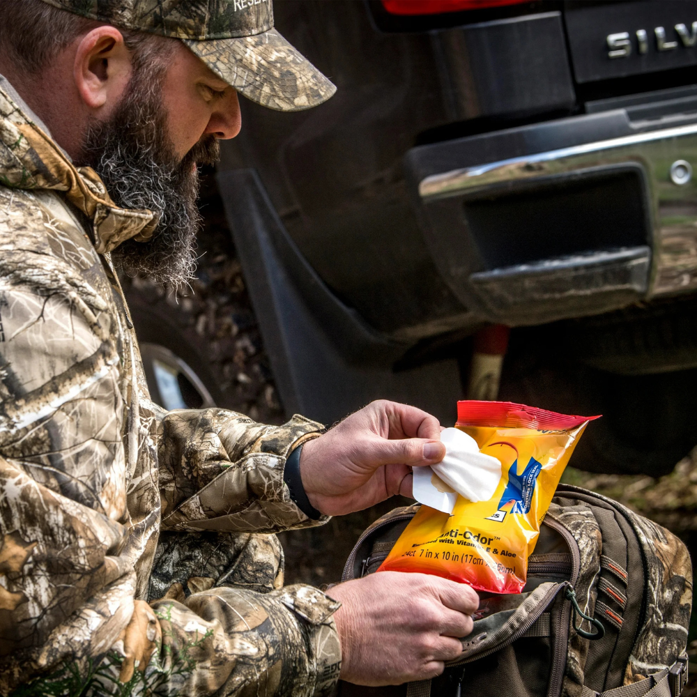 Person wearing camouflage clothing removing a scent-free wipe from Scent Killer Gold field wipes pack beside a backpack