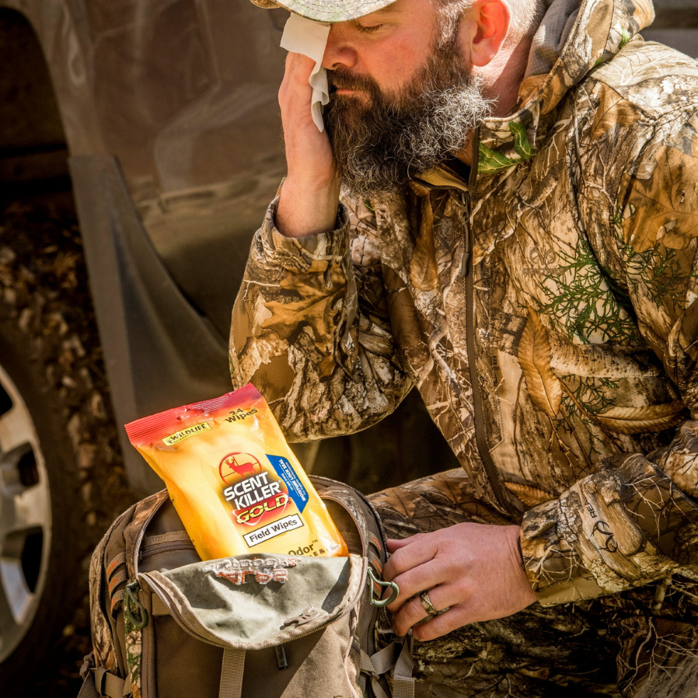 Person wearing camouflage clothing wiping face with scent-free wipe beside backpack containing Scent Killer Gold field wipes