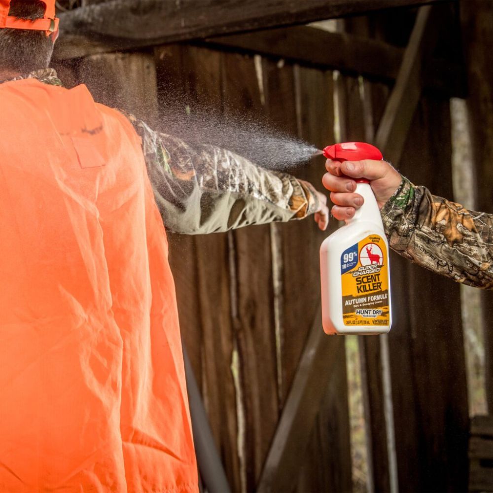 Person in camouflage holding a bottle of Scent Killer spray towards another person in an orange vest.