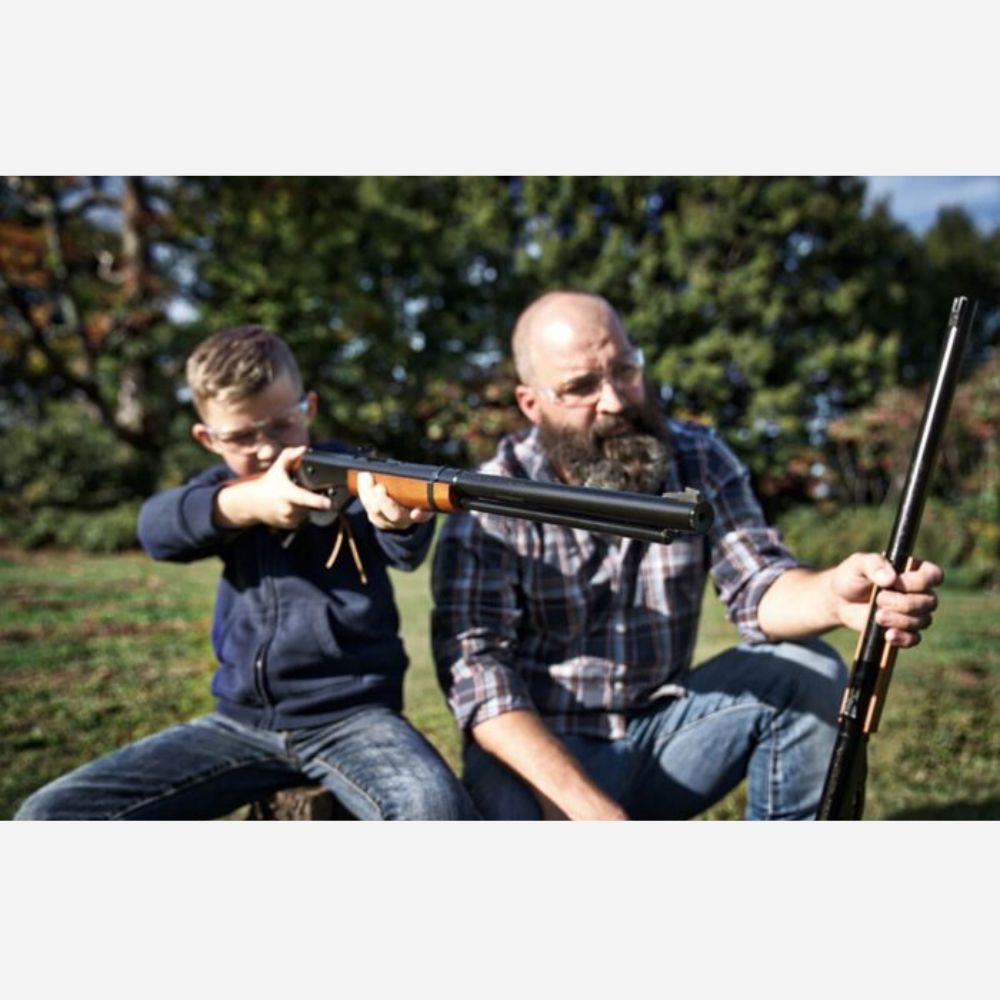 Man and child holding rifles outdoors with trees in the background
