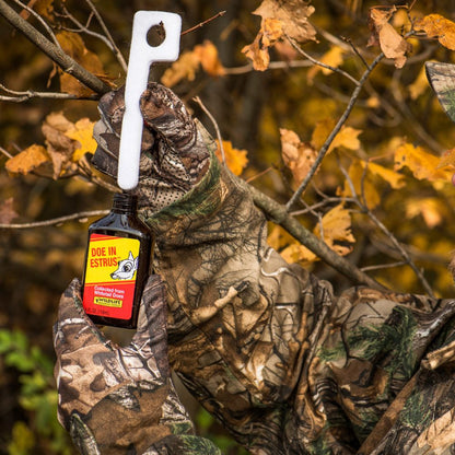 Person in camouflage holding a bottle of Doe in Estrus deer attractant with a blurred autumn background.