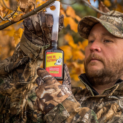 Man in camouflage holding a bottle of Doe In Estrus lure in an autumn forest setting