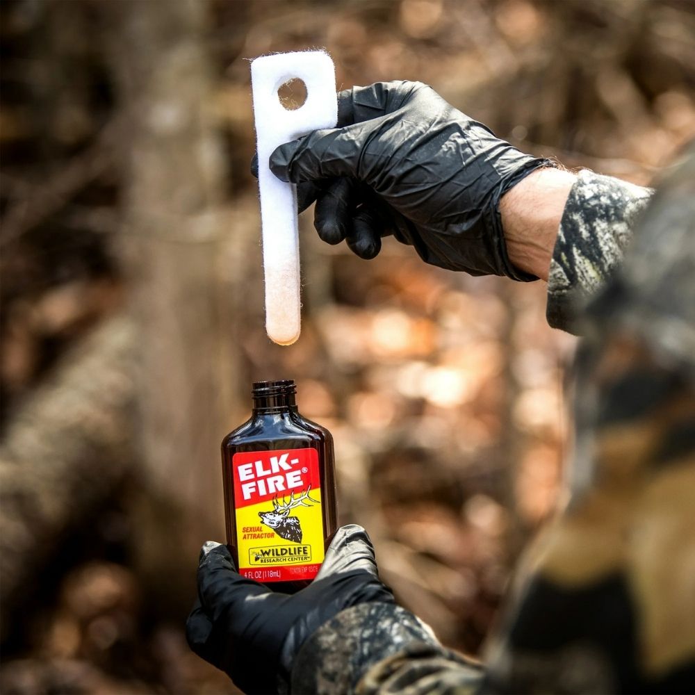 Gloved hands holding Elk-Fire scent attractant bottle and lifting a scent wick outdoors