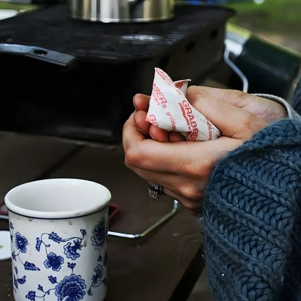 Person holding two tea bags over a floral mug on a camping stove.