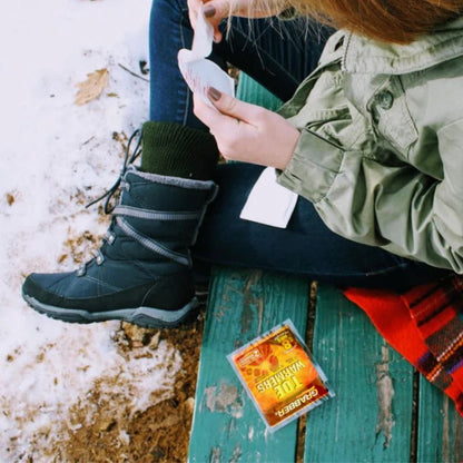 Person sitting on a bench with a snow-covered ground, wearing a green jacket and black boots.