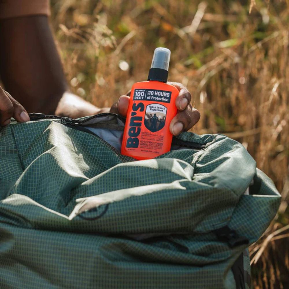Person holding a bottle of Ben's insect repellent over a green backpack in a natural setting.