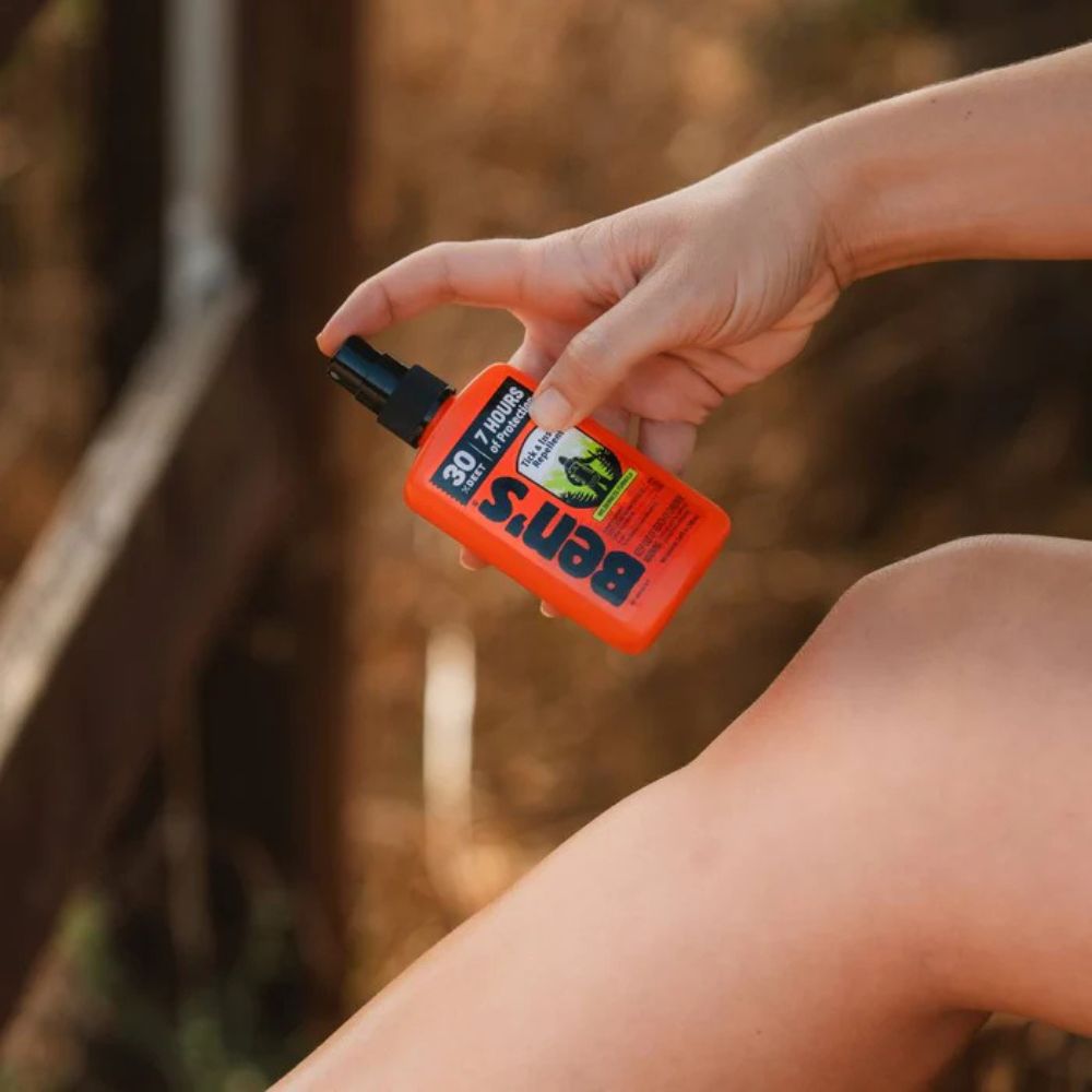 Hand holding an orange Ben's 6 Insect Repellent spray bottle against a blurred natural background