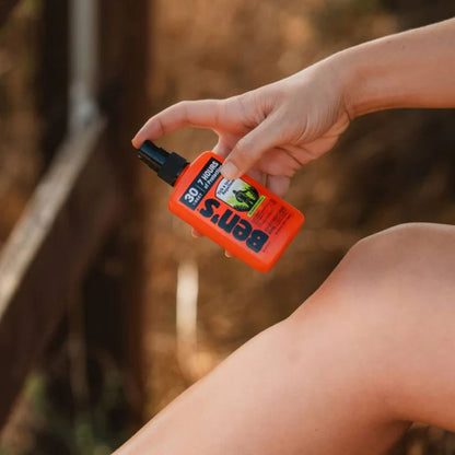 Hand holding an orange Ben's 6 Insect Repellent spray bottle against a blurred natural background