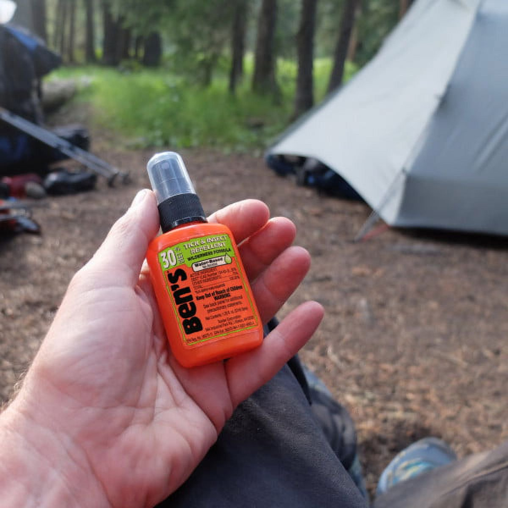 Hand holding an orange bottle of Ben's insect repellent with a camping scene in the background