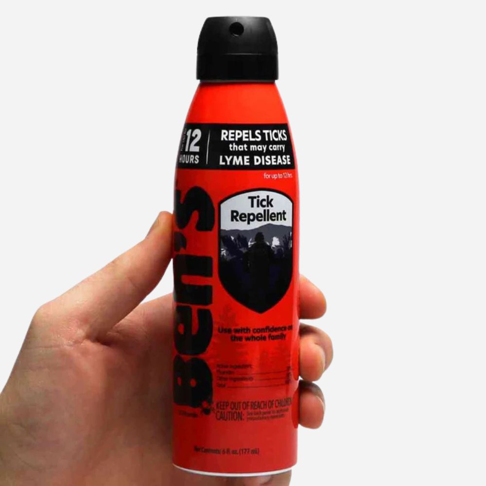 Hand holding a red and black tick repellent spray can against a white background