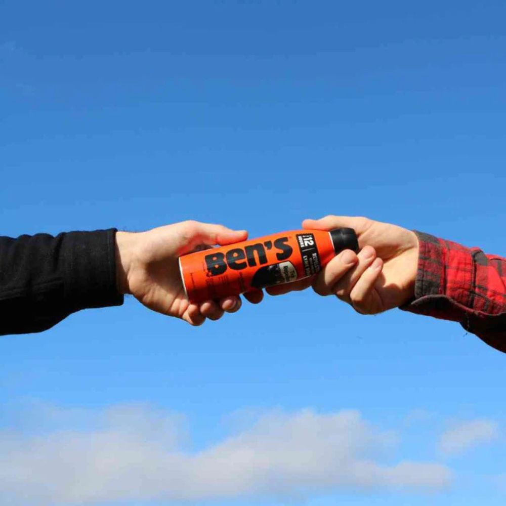 Two hands exchanging a can of Ben's insect repellent against a blue sky.