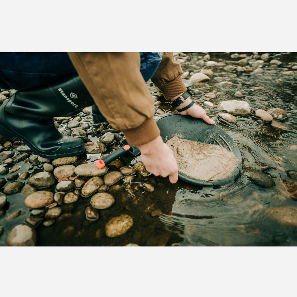 Person wearing rubber boots and a brown jacket holding a metal detector over water and rocks.