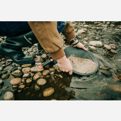 Person wearing rubber boots and a brown jacket holding a metal detector over water and rocks.