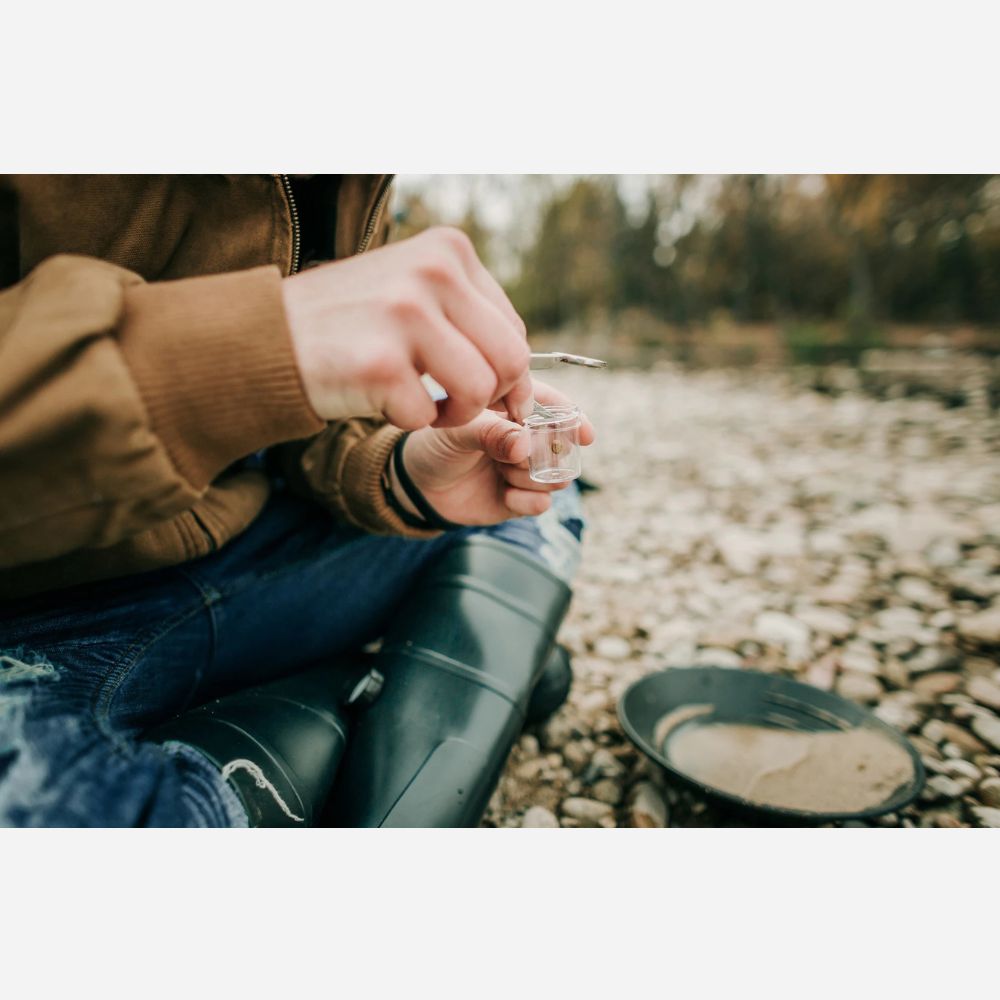 Person sitting outdoors by a stream, holding a small container.